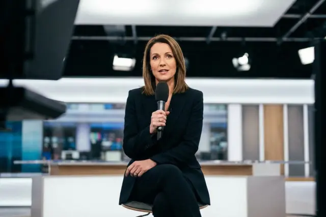 A female television presenter sitting in a bright broadcast studio holding a microphone